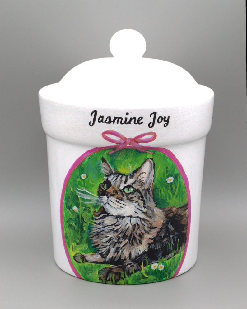 May include: White ceramic container with a lid, featuring a hand-painted portrait of a cat within a green and pink oval. The name "Jasmine Joy" is written above the cat portrait. A pink bow is below the name.