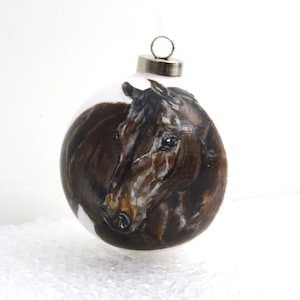 Pode incluir: Um enfeite de vidro pintado &agrave; m&atilde;o com a cabe&ccedil;a de um cavalo castanho e branco. O enfeite &eacute; redondo e tem um gancho prateado.