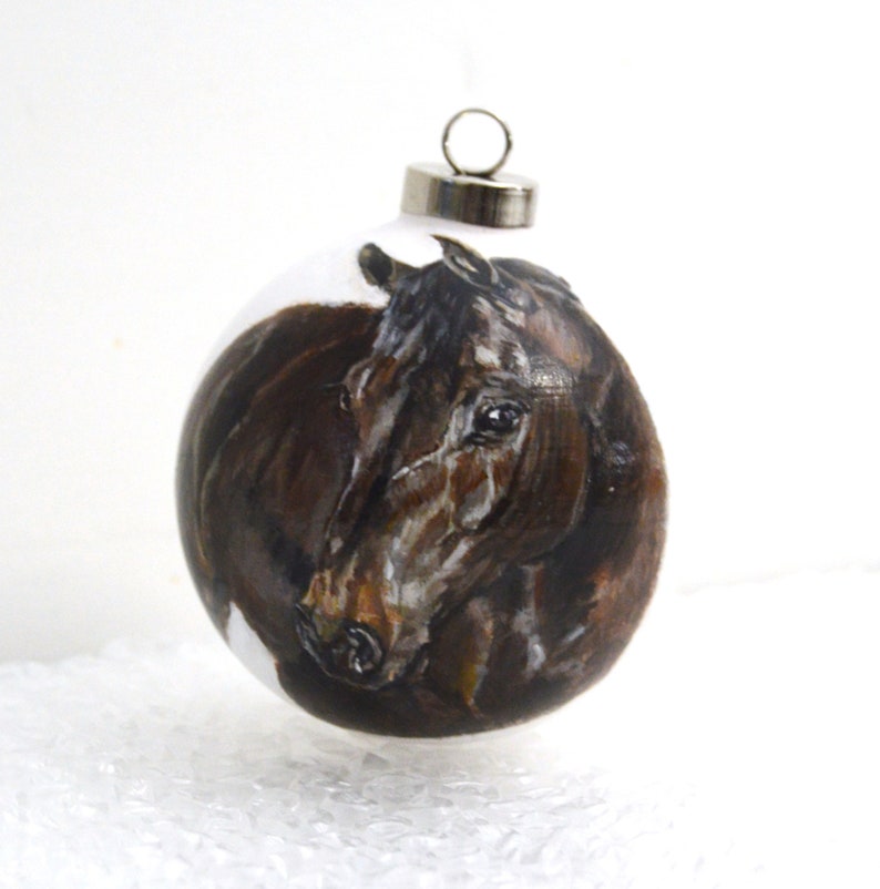 Pode incluir: Um enfeite de vidro pintado &agrave; m&atilde;o com a cabe&ccedil;a de um cavalo castanho e branco. O enfeite &eacute; redondo e tem um gancho prateado.
