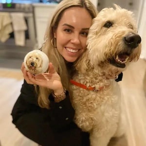 Pode incluir: Uma mulher est&aacute; segurando um enfeite de Natal branco com o rosto de um cachorro. Ela est&aacute; sorrindo e olhando para a c&acirc;mera. Um cachorro grande de cor marrom claro est&aacute; sentado ao lado dela e olhando para a c&acirc;mera.