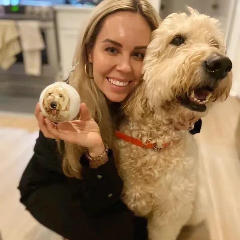 Pode incluir: Uma mulher est&aacute; segurando um enfeite de Natal branco com o rosto de um cachorro. Ela est&aacute; sorrindo e olhando para a c&acirc;mera. Um cachorro grande de cor marrom claro est&aacute; sentado ao lado dela e olhando para a c&acirc;mera.