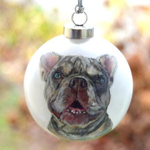 Pode incluir: Uma bola de Natal branca com uma imagem pintada &agrave; m&atilde;o de um Bulldogue Franc&ecirc;s. O c&atilde;o tem uma pelagem castanho claro e cinza com um peito branco e um nariz preto. Os olhos do c&atilde;o s&atilde;o azuis e ele tem a l&iacute;ngua rosa de fora.