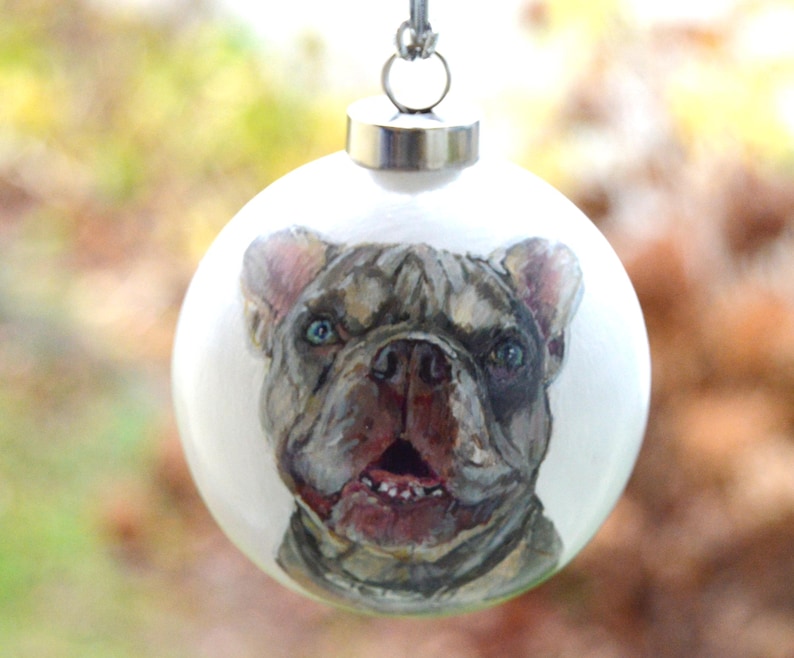 Pode incluir: Uma bola de Natal branca com uma imagem pintada &agrave; m&atilde;o de um Bulldogue Franc&ecirc;s. O c&atilde;o tem uma pelagem castanho claro e cinza com um peito branco e um nariz preto. Os olhos do c&atilde;o s&atilde;o azuis e ele tem a l&iacute;ngua rosa de fora.