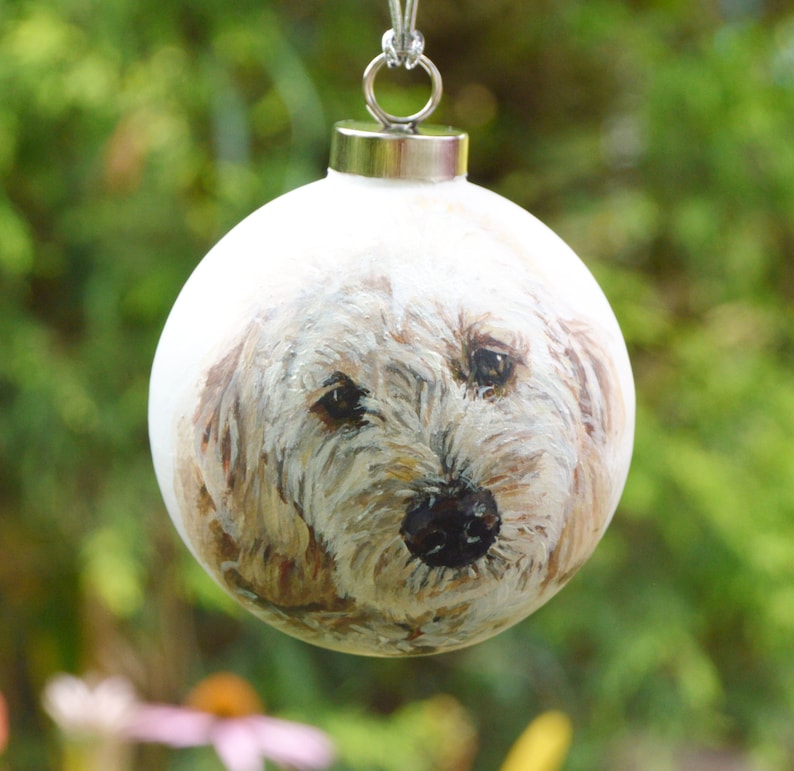 May include: A white Christmas ornament with a hand-painted portrait of a dog. The dog has brown and white fur and is looking directly at the viewer.
