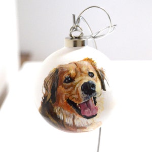 Pode incluir: Um enfeite de Natal branco com um retrato pintado &agrave; m&atilde;o de um c&atilde;o golden retriever. O c&atilde;o est&aacute; sorrindo com a l&iacute;ngua para fora.