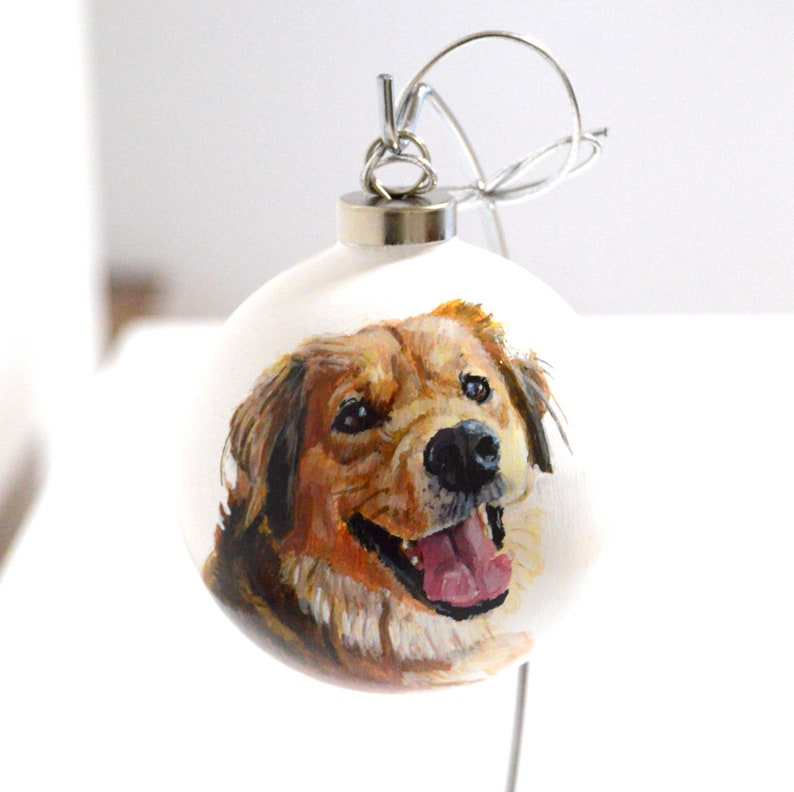 Pode incluir: Um enfeite de Natal branco com um retrato pintado &agrave; m&atilde;o de um c&atilde;o golden retriever. O c&atilde;o est&aacute; sorrindo com a l&iacute;ngua para fora.