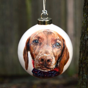 Pode incluir: Uma bola de Natal branca com um retrato pintado &agrave; m&atilde;o de um c&atilde;o castanho e branco com uma gravata borboleta vermelha e preta.