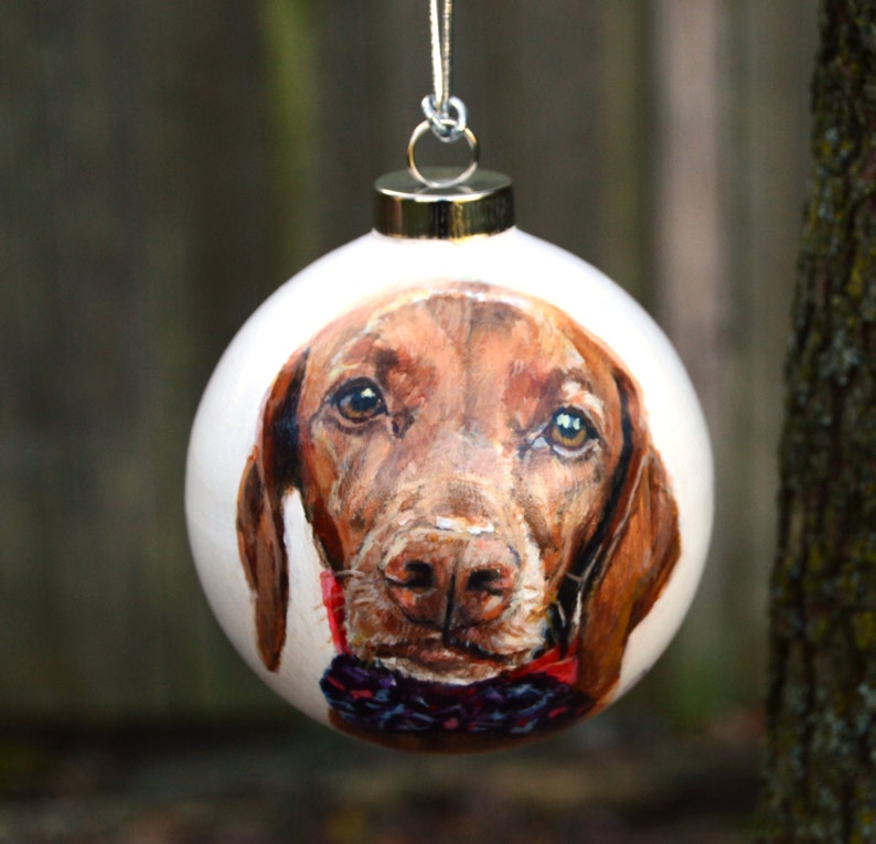 Pode incluir: Uma bola de Natal branca com um retrato pintado &agrave; m&atilde;o de um c&atilde;o castanho e branco com uma gravata borboleta vermelha e preta.