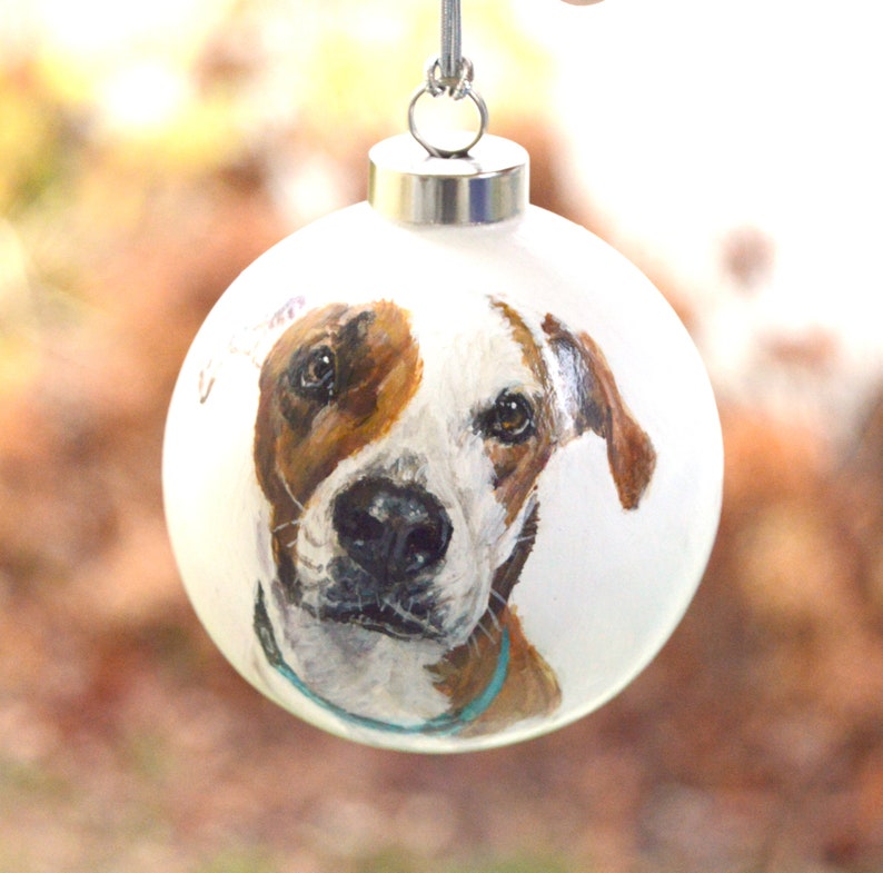 Pode incluir: Uma bola de Natal branca com um retrato pintado &agrave; m&atilde;o de um c&atilde;o castanho e branco. O c&atilde;o est&aacute; a olhar diretamente para o observador com uma express&atilde;o amig&aacute;vel. A bola tem um topo prateado e uma coleira azul turquesa.