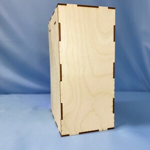 May include: A plain, light brown wooden box with a smooth surface and a slightly textured grain. The box has a simple, rectangular shape and is made of plywood.