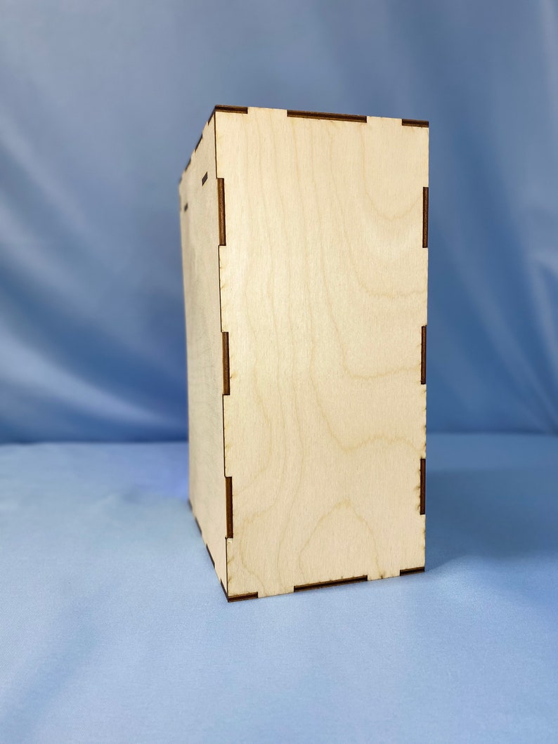 May include: A plain, light brown wooden box with a smooth surface and a slightly textured grain. The box has a simple, rectangular shape and is made of plywood.