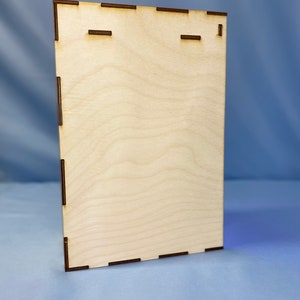 May include: A plain, light brown wooden plaque with a rectangular shape and a smooth surface. The plaque has two small, rectangular cutouts near the top.