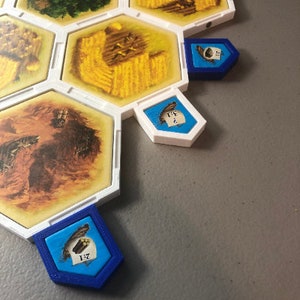 May include: A close-up of a board game with hexagonal tiles. The tiles are white with brown and yellow illustrations. There are blue and white tokens with numbers on them.