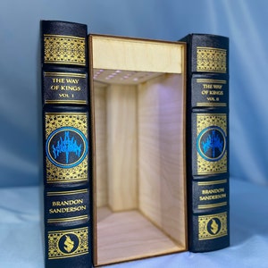 May include: Two black hardback books with gold lettering and decorative designs. The books are titled "The Way of Kings" Vol. I and Vol. II by Brandon Sanderson. The books are open to reveal a wooden box with a light inside.