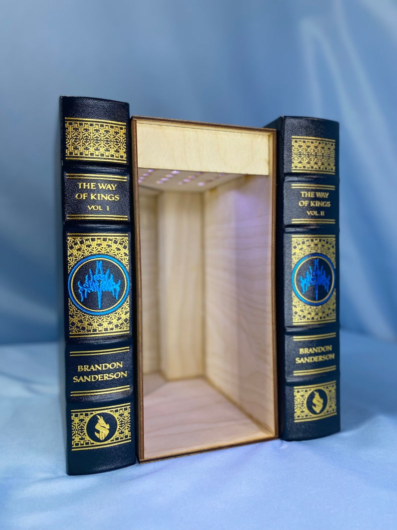 May include: Two black hardback books with gold lettering and decorative designs. The books are titled "The Way of Kings" Vol. I and Vol. II by Brandon Sanderson. The books are open to reveal a wooden box with a light inside.