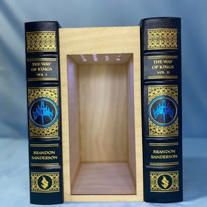 May include: Two black leather-bound books with gold lettering and illustrations. The books are titled "The Way of Kings" Vol. 1 and Vol. 2 by Brandon Sanderson. The books are displayed in a wooden bookend with a light inside.