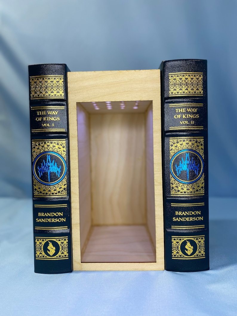 May include: Two black leather-bound books with gold lettering and illustrations. The books are titled "The Way of Kings" Vol. 1 and Vol. 2 by Brandon Sanderson. The books are displayed in a wooden bookend with a light inside.