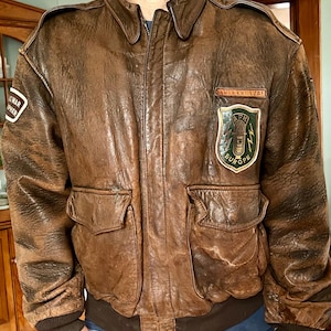 May include: A brown leather bomber jacket with a distressed finish. The jacket features a zip-up front, two front pockets, and patches on the chest and sleeve. The patches include text and a green and yellow emblem. The jacket has a ribbed collar and cuffs.
