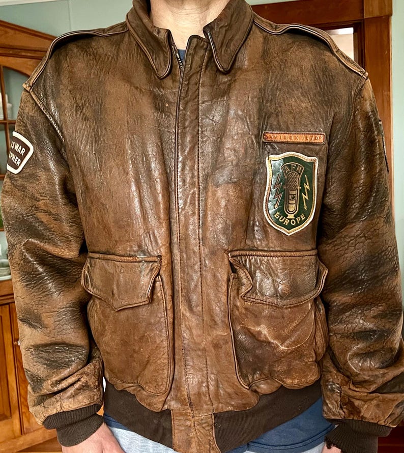 May include: A brown leather bomber jacket with a distressed finish. The jacket features a zip-up front, two front pockets, and patches on the chest and sleeve. The patches include text and a green and yellow emblem. The jacket has a ribbed collar and cuffs.