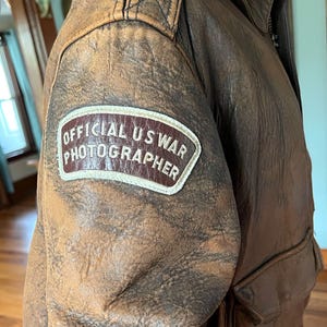 May include: A vintage brown leather jacket with a distressed appearance. The jacket features a patch on the upper arm that reads "OFFICIAL US WAR PHOTOGRAPHER". The jacket has a zipper closure and a pocket.