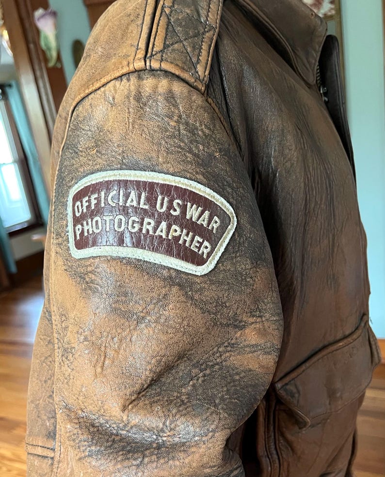 May include: A vintage brown leather jacket with a distressed appearance. The jacket features a patch on the upper arm that reads "OFFICIAL US WAR PHOTOGRAPHER". The jacket has a zipper closure and a pocket.
