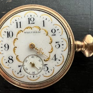 May include: A gold-tone Waltham pocket watch with a white dial, black Arabic numerals, and ornate gold filigree detailing surrounding the center. The watch features a small sub-dial for seconds at the bottom and a winding crown on the right side.