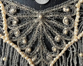 Può includere: Una collana vintage di perline con un design delicato e intricato. La collana presenta un motivo centrale a forma di ventaglio con una frangia di perline che pendono. Le perline sono un mix di argento e bianco, creando un effetto scintillante.