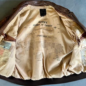May include: Brown leather bomber jacket with a tan lining. The lining features text that reads "AVIREX FLYING TIGERS" and a diagram. The jacket has multiple pockets and patches. The jacket is open, displaying the interior.