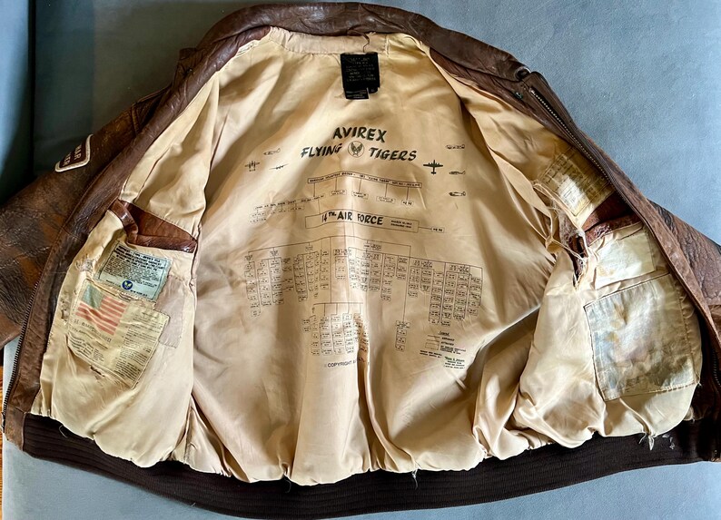 May include: Brown leather bomber jacket with a tan lining. The lining features text that reads "AVIREX FLYING TIGERS" and a diagram. The jacket has multiple pockets and patches. The jacket is open, displaying the interior.