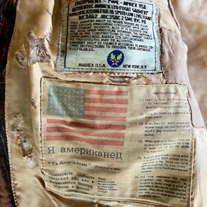 May include: Close-up of the interior of a vintage brown leather flight jacket. The lining features a sewn-in label with an American flag and text in English and Russian. Additional labels provide instructions and specifications.