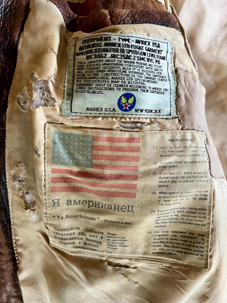 May include: Close-up of the interior of a vintage brown leather flight jacket. The lining features a sewn-in label with an American flag and text in English and Russian. Additional labels provide instructions and specifications.