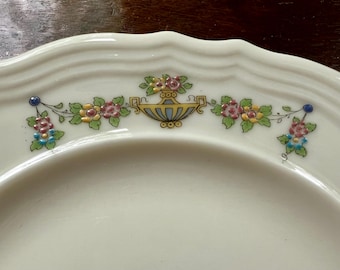 May include: Cream-colored ceramic plate with a scalloped edge. The plate features a floral garland design in pink, yellow, blue, and green. A central vase design is also present, adding to the decorative appeal. The plate is likely for serving food.
