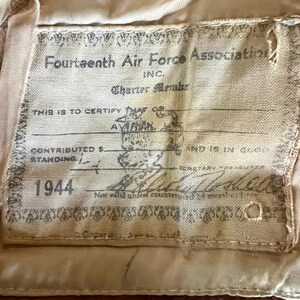 May include: A tan fabric label with the text "Fourteenth Air Force Association" and "1944". The label includes handwritten signatures and stamps, with the text "THIS IS TO CERTIFY" and "AVIRAK".