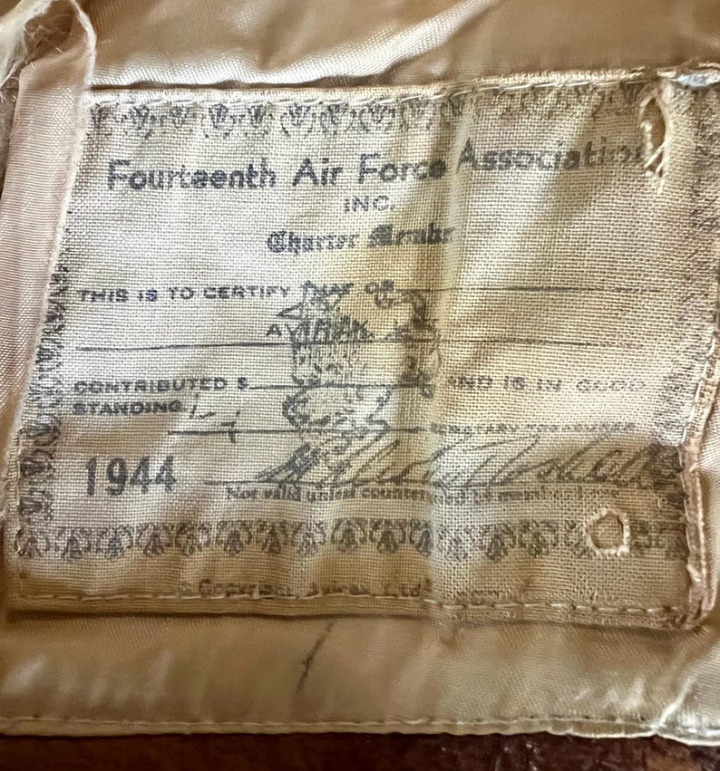 May include: A tan fabric label with the text "Fourteenth Air Force Association" and "1944". The label includes handwritten signatures and stamps, with the text "THIS IS TO CERTIFY" and "AVIRAK".