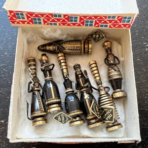 May include: A collection of vintage black and gold decorative bottle stoppers in a box. The stoppers have various shapes and designs, including figures and geometric patterns. Each stopper has a small tag that reads "TEPPICH 1930".