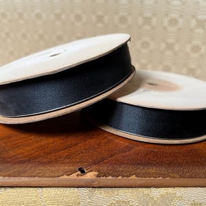May include: Two spools of black satin ribbon with a thin white border. The ribbon is wound around a light brown cardboard spool. The spools are on a dark wooden surface.