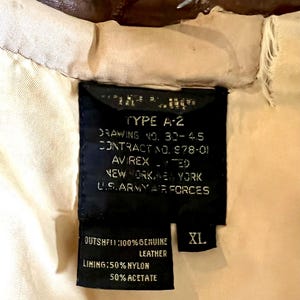 May include: Close-up of a black label from an A-2 jacket, with white text indicating the jacket's specifications. The label reads "TYPE A-2" and "U.S. ARMY AIR FORCES." The label also indicates the jacket is XL and made of genuine leather.