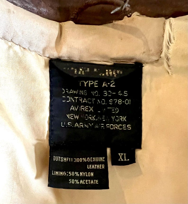 May include: Close-up of a black label from an A-2 jacket, with white text indicating the jacket's specifications. The label reads "TYPE A-2" and "U.S. ARMY AIR FORCES." The label also indicates the jacket is XL and made of genuine leather.