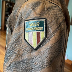 May include: Close-up of a brown leather jacket with a distressed finish. A patch on the jacket reads "WAR CORRESPONDENT" in white text on a green background, with a colorful emblem below. The jacket has a vintage, worn appearance.