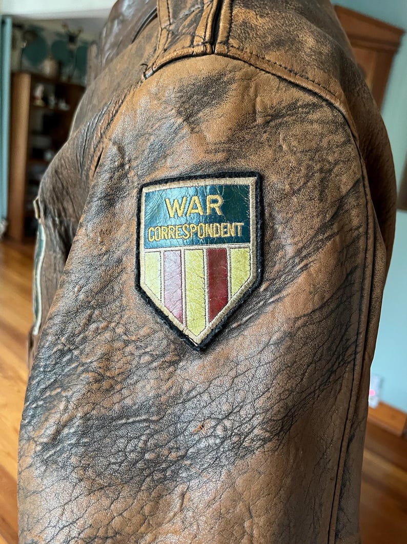 May include: Close-up of a brown leather jacket with a distressed finish. A patch on the jacket reads "WAR CORRESPONDENT" in white text on a green background, with a colorful emblem below. The jacket has a vintage, worn appearance.