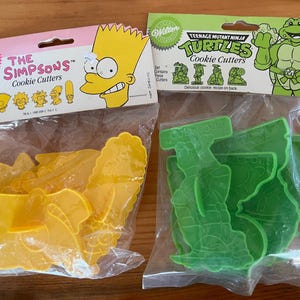 May include: Two sets of cookie cutters in clear plastic packaging. One set is yellow and features characters from "The Simpsons." The other set is green and features characters from "Teenage Mutant Ninja Turtles." Both sets are from Wilton.