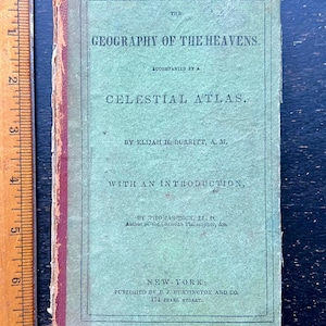 May include: An antique book titled "The Geography of the Heavens" with "Celestial Atlas" on the cover. The cover is a faded green with red trim and text in black. The book is next to a wooden ruler.