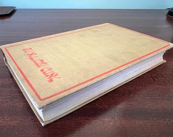May include: A vintage book with a tan cover and red border. The book's title is printed in red Hebrew lettering. The pages are white and the book is resting on a dark brown wooden surface.
