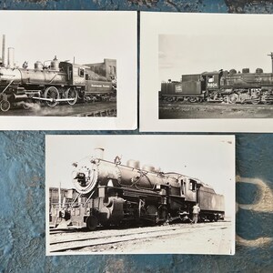May include: Three black and white photographs of steam locomotives. The images show the trains from different angles, highlighting their intricate details. The locomotives are on tracks, with some smoke visible, suggesting they are in operation. The photographs are on white paper.