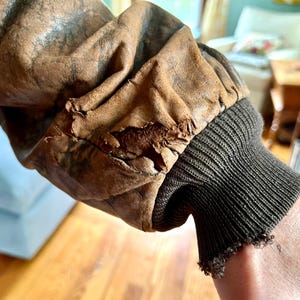 May include: Close-up of a vintage brown leather jacket sleeve. The leather shows wear and tear, with visible cracks and distressed areas. The sleeve features a dark green ribbed knit cuff, adding a contrasting texture to the aged leather.