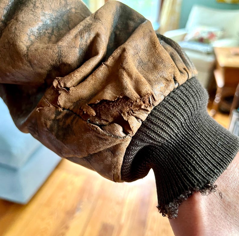 May include: Close-up of a vintage brown leather jacket sleeve. The leather shows wear and tear, with visible cracks and distressed areas. The sleeve features a dark green ribbed knit cuff, adding a contrasting texture to the aged leather.