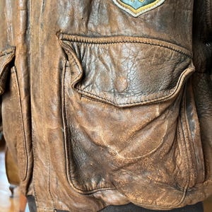 May include: A distressed brown leather bomber jacket with multiple pockets and a ribbed hem. A patch with the word "EUROPE" is visible on the upper left shoulder. The jacket has a worn, vintage appearance.