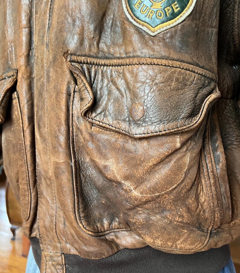 May include: A distressed brown leather bomber jacket with multiple pockets and a ribbed hem. A patch with the word "EUROPE" is visible on the upper left shoulder. The jacket has a worn, vintage appearance.