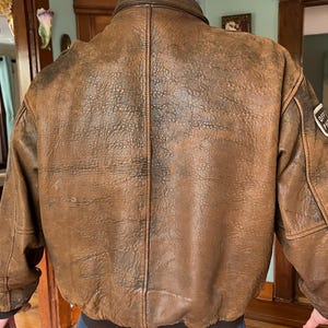 May include: A brown leather bomber jacket with a distressed finish. The jacket features a ribbed hem and cuffs, a zip-up front, and a collar. The back of the jacket is visible, showing the texture of the leather and the stitching details. A patch is visible on the upper arm.