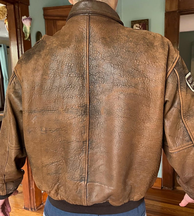 May include: A brown leather bomber jacket with a distressed finish. The jacket features a ribbed hem and cuffs, a zip-up front, and a collar. The back of the jacket is visible, showing the texture of the leather and the stitching details. A patch is visible on the upper arm.
